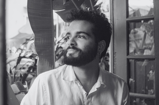 Retrato em preto e branco de Wallacy Coelho, arquiteto, em janela com folhagem ao fundo.