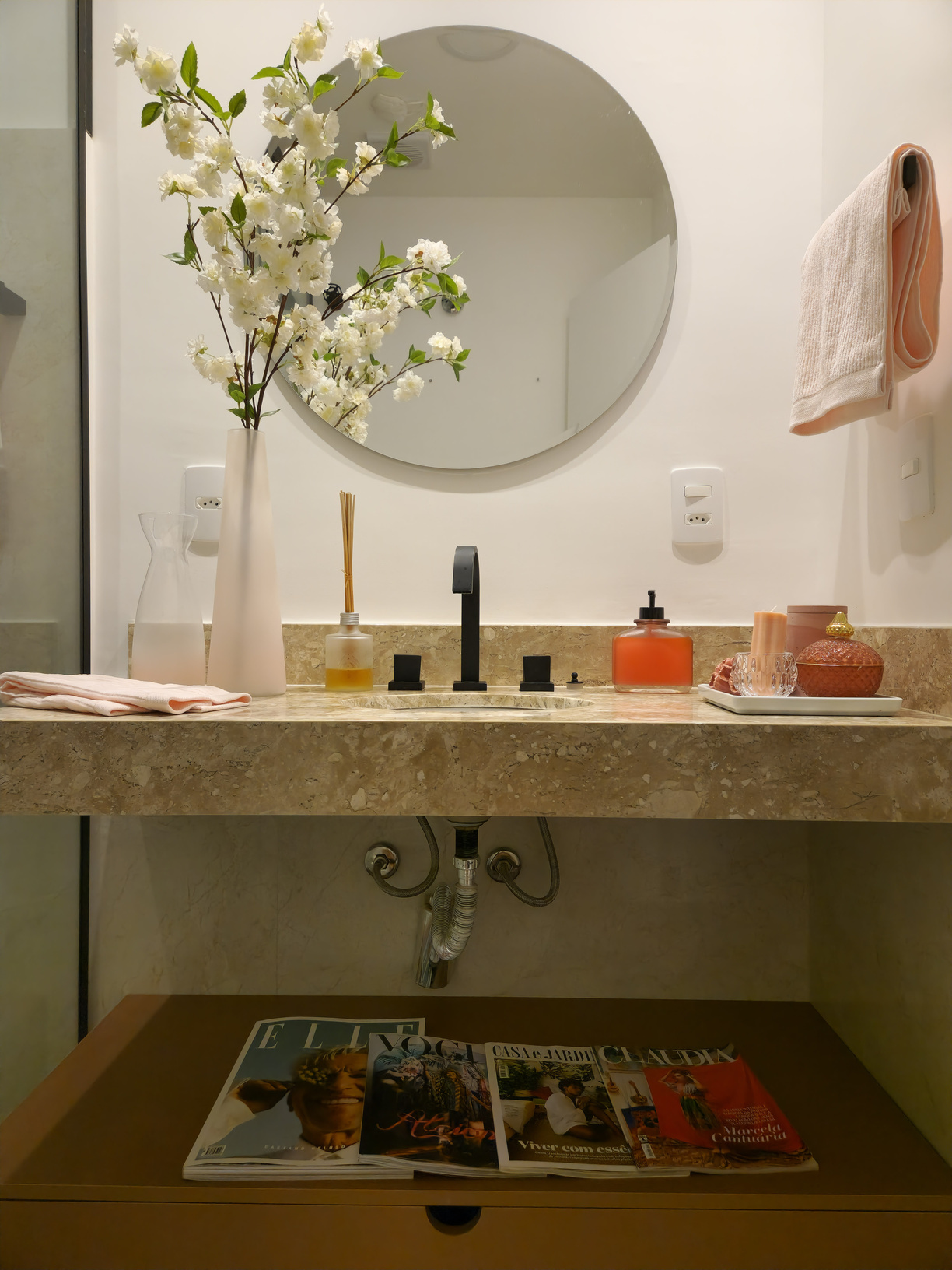 Lavabo com cuba esculpida em mármore, espelho redondo, flores brancas e revistas dispostas na prateleira inferior.