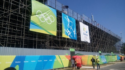 Arquibancada com estrutura metálica vista do nível da rua, com bandeiras dos Jogos Rio 2016 e equipe de obra ao redor.