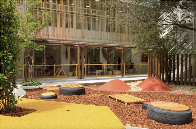 Pátio externo da escola com tanques de areia, círculos de troncos de madeira e a fachada amarela canelada ao fundo.