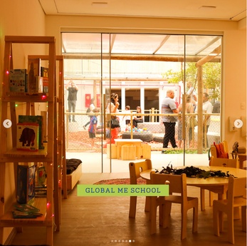 Entrada da Global Me School vista do interior, com estante de livros e brinquedos e crianças no pátio externo.