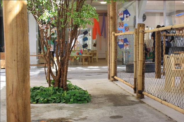 Pátio interno de escola infantil com árvore central, piso de cimento, divisórias de bambu e tela aramada, banco de madeira ao fundo.