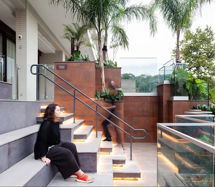 Arquibancadas-jardim que conectam dois desníveis, com jardineiras suspensas em corten e mulher sentada nos degraus.