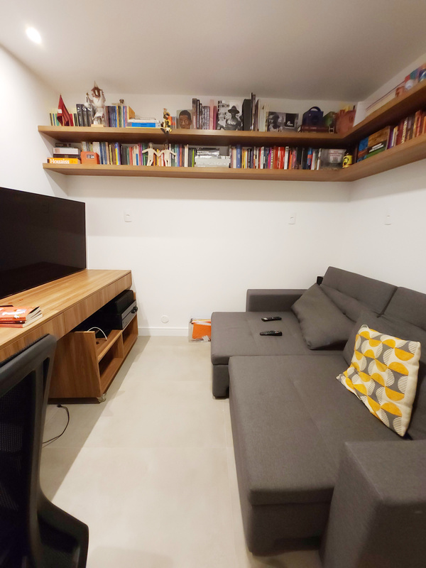 Sala de TV com biblioteca em L no alto da parede, sofá chaise cinza, almofada estampada e tapete claro.