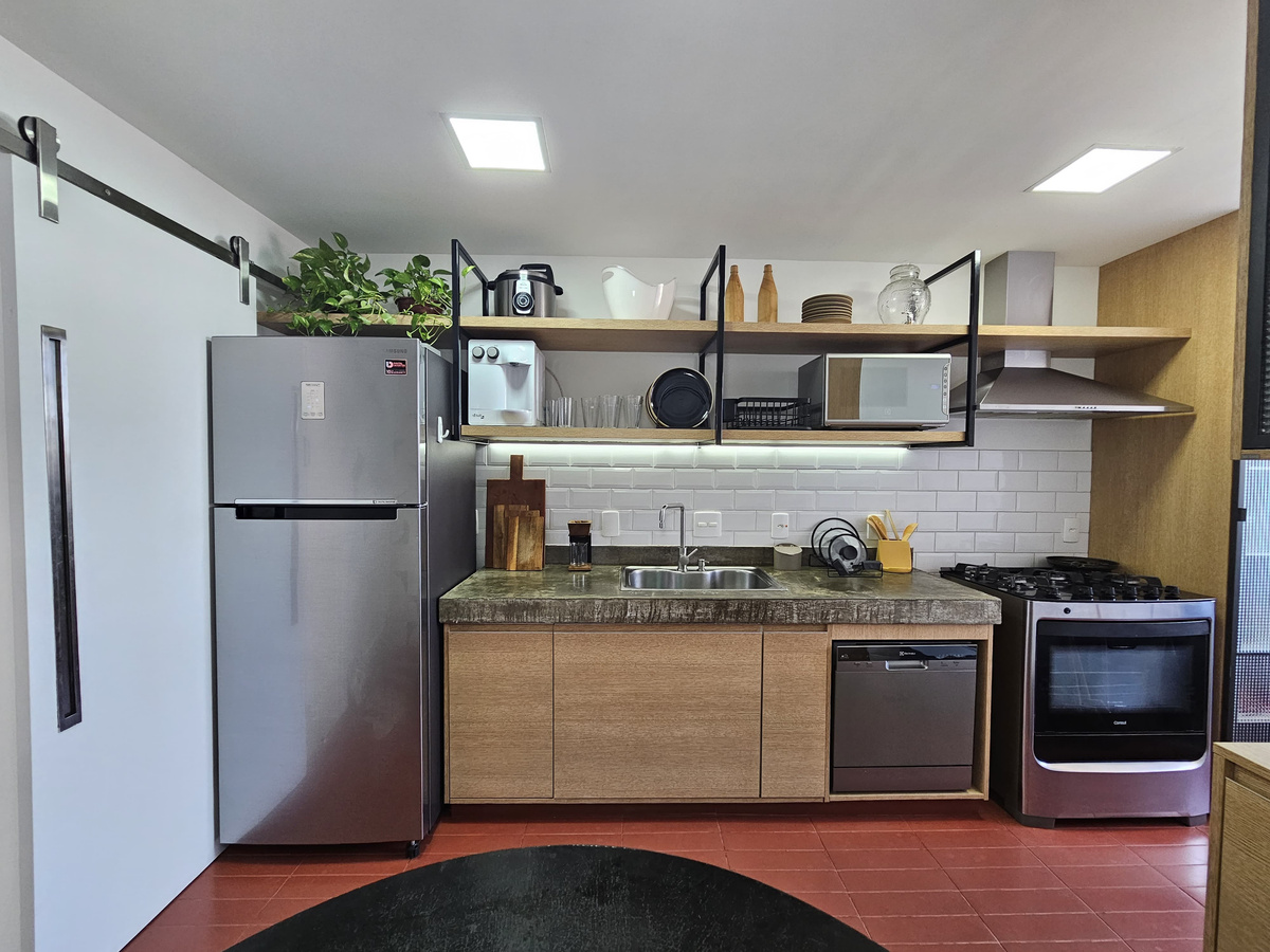 Cozinha residencial com piso terracota, prateleiras metálicas suspensas, geladeira e fogão de aço inox, parede branca com subway tiles.