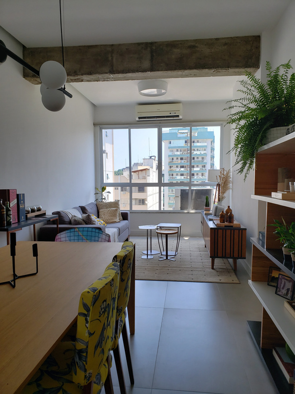 Sala de jantar e estar vista do canto, com mesa de madeira, cadeiras de chita amarela, janela ampla com vista para a cidade e estante na parede oposta.