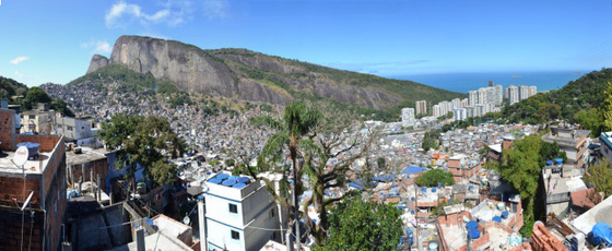 Vista panorâmica da Rocinha com a Pedra Dois Irmãos à esquerda e o mar de São Conrado à direita, telhados se acumulando até a base do morro.